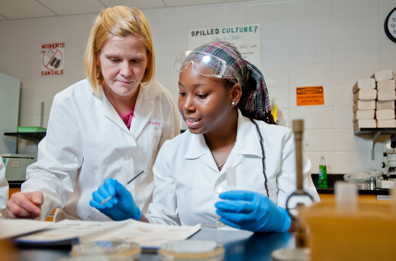 A teacher in a white lab coat instructs a student in a lab setting